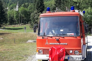 Frontansicht eines rotfarbenen Feuerwehrfahrzeugs der Marke Magirus Deutz mit Blaulichtern auf dem Dach, geparkt auf einem Weg in einer grünen, waldreichen Landschaft. | © Technische Sammlung Haus Schleusenfleth 