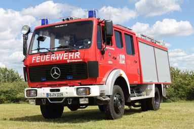 Rotes Feuerwehrfahrzeug (Mercedes-Benz) mit Blaulicht und Aufschrift ‚Feuerwehr‘, geparkt auf einer Wiese unter blauem Himmel. | © Feuerwehr Nidda 