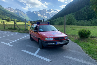 Rotes Feuerwehrfahrzeug mit Blaulicht steht auf einem Parkplatz in einem alpinen Tal; im Hintergrund grüne Wiesen, bewaldete Hänge und schneebedeckte Berge unter blauem Himmel. | © Feuerwehr Wolpertshausen 