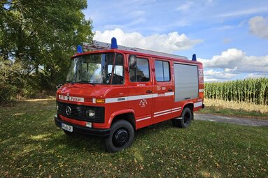 Rotes Feuerwehrfahrzeug (Mercedes-Benz) mit Blaulicht steht auf einer Wiese neben einem Feld; im Hintergrund Bäume und ein Maisfeld unter blauem Himmel mit Wolken. | © Löschgruppe Putzar 
