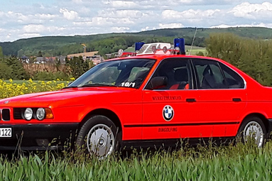 Rotes BMW-Einsatzfahrzeug der Feuerwehr mit Blaulicht auf dem Dach, geparkt am Rand eines Feldes mit Hügellandschaft im Hintergrund. | © Bernd Donsbach 