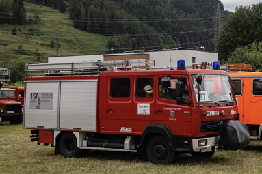 Ein rotes Feuerwehrfahrzeug steht auf einer Wiese vor einem Gebäude. Auf dem Dach sind Leitern befestigt, oben befinden sich blaue Rundumleuchten. An der Seite ist „Freiwillige Feuerwehr“ zu sehen. Im Hintergrund befinden sich bewaldete Berge. | © Freiwillige Feuerwehr Söcking e.V. 