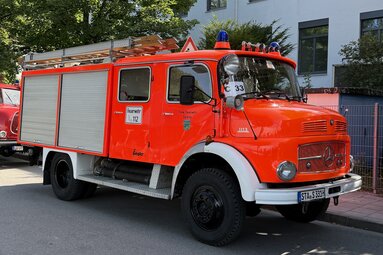 Ein rotes Feuerwehrfahrzeug (älteres Modell) steht am Straßenrand. Es hat silberne Rollläden an den Seiten, eine ausziehbare Leiter auf dem Dach und blaue Rundumleuchten. Auf der Fahrertür ist „Feuerwehr“ zu lesen. Im Hintergrund sind ein Gebäude und Bäume zu sehen. | © Freiwillige Feuerwehr Söcking e.V.