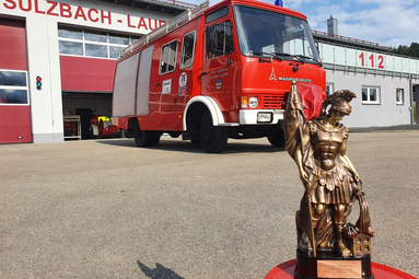 Bronzefarbene Statue im Vordergrund; dahinter ein rotes Feuerwehrfahrzeug vor einem Feuerwehrhaus mit der Aufschrift ‚Sulzbach-Laufen‘ und der Notrufnummer 112. | © Feuerwehr Sulzbach-Laufen 