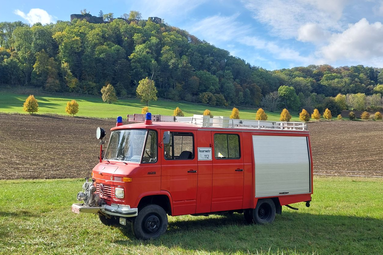 Rotes Feuerwehrfahrzeug mit Blaulicht steht auf einer Wiese vor Feldern und bewaldetem Hügel unter blauem Himmel. | © Thomas Herr 