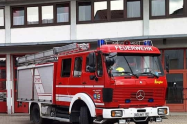 Rotes Feuerwehr-Löschfahrzeug mit der Aufschrift „FEUERWEHR“ auf der Front, vor einem Feuerwehrgebäude geparkt; auf dem Dach sind Leitern befestigt, blaue Rundumleuchten und seitliche Geräteraumklappen sichtbar. | © Freiwillige Feuerwehr Wiesloch 