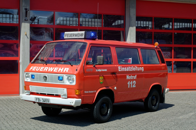 Ein roter Feuerwehr-Kleinbus mit der Aufschrift „FEUERWEHR“, „Einsatzleitung“ und „Notruf 112“ steht vor einer Feuerwache mit roten Hallentoren. Auf dem Dach befinden sich Blaulicht und eine Leuchttafel mit der Aufschrift „Feuerwehr“. | © Luca Blecher 