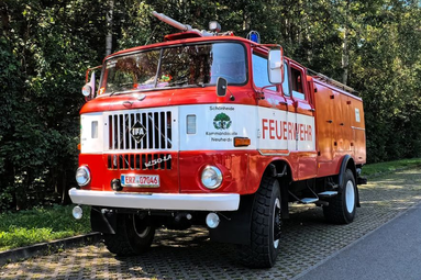Rotes historisches Feuerwehrfahrzeug mit weißer Front und der Aufschrift ‚Feuerwehr‘, geparkt am Straßenrand vor einem Wald. | © Freiwillige Feuerwehr Schönheide 