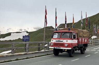 Alter roter Feuerwehr-LKW auf einer Hochalpenstraße vor Bergkulisse; neben der Fahrbahn stehen mehrere Fahnen und ein Schild mit der Aufschrift „Parkplatz Fuscher Törl“. | © Historikabteilung der Feuerwehr Breitenau-NL