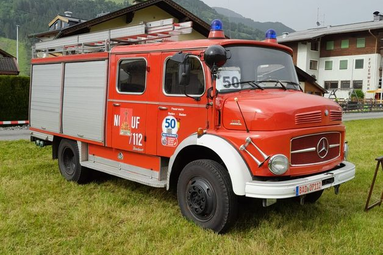 Rotes Feuerwehrfahrzeug (älteres Modell) mit seitlichen Rollläden und Blaulichtern auf dem Dach, geparkt auf einer Wiese vor Häusern und Bergen im Hintergrund. | © Historischer Feuerwehr- und Kulturverein Baden Baden e.V.