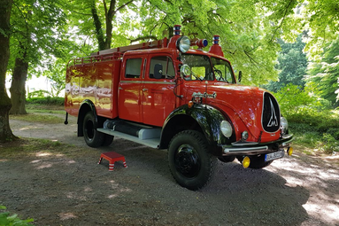 Historisches rotes Feuerwehrfahrzeug mit Blaulicht steht auf einem Waldweg unter grünen Bäumen. | © Ulrich Bauer 