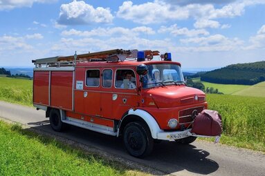 Historisches rotes Feuerwehrfahrzeug auf einer ländlichen Straße, umgeben von grünen Wiesen unter blauem Himmel mit weißen Wolken. | © Feuerwehr Engen 