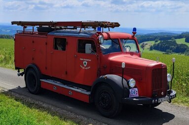 Historisches rotes Feuerwehrfahrzeug auf einer schmalen Landstraße in hügeliger Landschaft, mit Leitern auf dem Dach und weitem Blick über grüne Felder. | © Feuerwehr Engen 