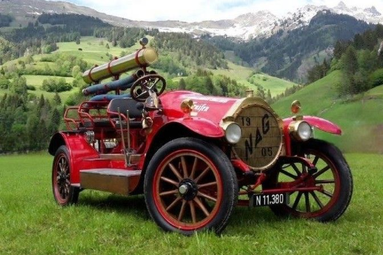 Historisches rotes Feuerwehrfahrzeug mit Holzspeichenrädern und montierter Feuerlöschpumpe auf einer grünen Wiese vor alpiner Berglandschaft. | © BTF VÖEST und Georg Fischer Traisen