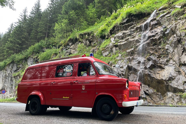Historisches rotes Feuerwehrfahrzeug auf einer Bergstraße, vor felsiger Kulisse mit kleinem Wasserfall und bewaldetem Hang geparkt. | © Freiwillige Feuerwehr Lindach 