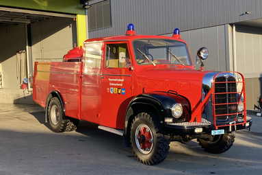 Historisches rotes Feuerwehrfahrzeug mit Blaulichtern und robustem Frontschutz, geparkt vor einem modernen Feuerwehrgebäude. | © Feuerwehrverein Embrachertal 