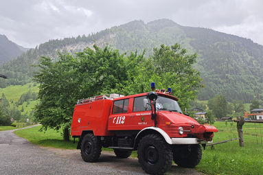 Rotes historisches Feuerwehr-Einsatzfahrzeug in alpiner Landschaft, geparkt auf einer Straße vor grünen Wiesen, Bäumen und bewaldeten Bergen. | © Freiwillige Feuerwehr Bad Tölz
