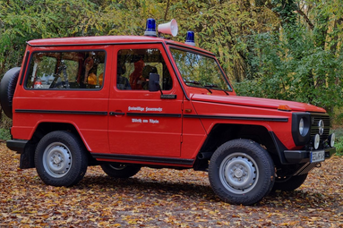 Roter historischer Feuerwehr-Geländewagen mit Blaulicht, geparkt auf herbstlichem Waldboden zwischen bunten Bäumen. | © Fabian Neumann 