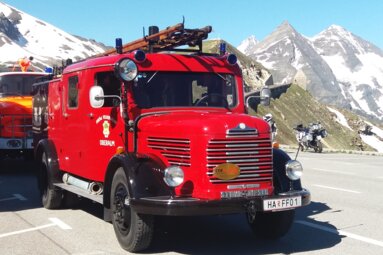 Historisches rotes Feuerwehrfahrzeug vor beeindruckender Hochgebirgskulisse mit schneebedeckten Bergen an einer Passstraße. | © Freiwillige Feuerwehr Oberalm