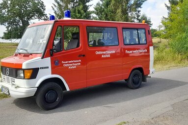 Roter historischer Feuerwehrbus mit Blaulichtern und Vereinsbeschriftung, geparkt auf einer Straße am Waldrand. | © Oldtimerfreunde Alsfeld