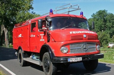 Historisches rotes Mercedes-Feuerwehrfahrzeug mit Blaulichtern und Dachleiter, unterwegs auf einer Straße in grüner Umgebung. | © Feuerwehr Oldtimer Verein Hard 