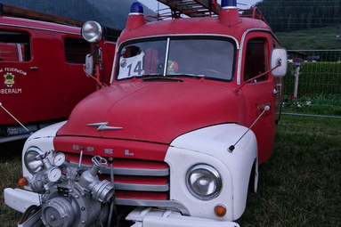 Historisches rotes Feuerwehrfahrzeug mit weißer Front und aufgebauter Leiter auf einer Wiese bei einer Oldtimerveranstaltung vor alpiner Bergkulisse. | © Roland Stohrer 