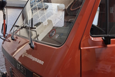 Detailaufnahme eines historischen Feuerwehrfahrzeugs in roter Lackierung. Sichtbar sind die Frontkabine mit großer Windschutzscheibe, Scheibenwischer, Seitenspiegel sowie die Beschriftung „Schlingmann“ und die Typenbezeichnung „LT 50“. Das Fahrzeug steht in einem Innenhof oder einer Fahrzeughalle. | © Helmut Dietz