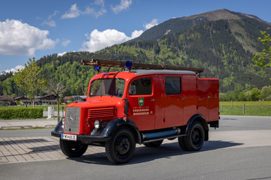 Historisches rotes Feuerwehrfahrzeug der Freiwilligen Feuerwehr Maishofen auf einem befestigten Platz vor einer grünen Berglandschaft. Das Oldtimer-Fahrzeug verfügt über eine Leiter und Blaulichter auf dem Dach. Im Hintergrund sind Wiesen, Bäume und bewaldete Berghänge unter einem sonnigen Himmel mit einzelnen Wolken zu sehen. | © Freiwillige Feuerwehr Maishofen 