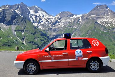 Rotes Feuerwehr-Einsatzfahrzeug auf der Großglockner Hochalpenstraße vor einer eindrucksvollen Hochgebirgskulisse. Im Hintergrund sind die schneebedeckten Gipfel des Großglockners und umliegende Berge zu sehen. Das Fahrzeug trägt die Beschriftung „Freiwillige Feuerwehr“ sowie eine Startnummer der Oldtimer Feuerwehr Weltmeisterschaft. Der Himmel ist klar und sonnig. | © Feuerwehr Bebra 