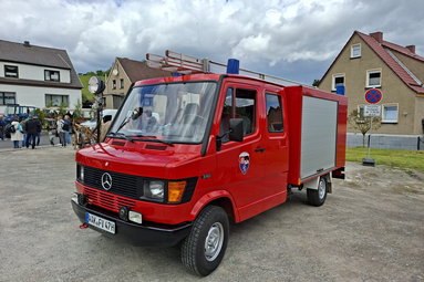 Rotes Feuerwehrfahrzeug der Marke Mercedes-Benz auf einem Festgelände bei einer Oldtimer-Veranstaltung. Das Fahrzeug steht auf einem Platz vor Wohnhäusern, im Hintergrund sind Besucherinnen und Besucher sowie ein bewölkter Himmel zu sehen. Das historische Einsatzfahrzeug ist mit Blaulicht und einer Leiter auf dem Dach ausgestattet. | © Eric Deisenroth 