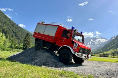 Historisches rotes Feuerwehrfahrzeug im alpinen Gelände, auf einem Schotterhügel positioniert, vor beeindruckender Bergkulisse mit Wäldern und schneebedeckten Gipfeln. | © Feuerwehr Meßstetten 