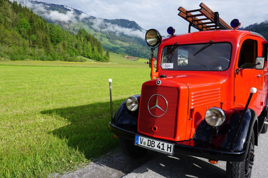 Historisches rotes Feuerwehrfahrzeug mit Leiteraufbau, auf einer Landstraße vor grüner Berglandschaft und bewaldeten Hängen geparkt. | © Traditionsverein Feuerwehr Oberheinsdorf e.V. 