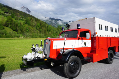 Historisches rot-weißes Feuerwehrfahrzeug mit offenem Aufbau und Frontpumpe, auf einer Landstraße vor alpiner Bergkulisse geparkt. | © Traditionsverein Feuerwehr Oberheinsdorf e.V.