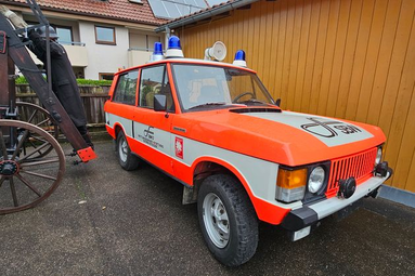 Historisches Feuerwehr-Kommandofahrzeug in orange-weißer Lackierung mit Blaulicht, vor einem Gebäude geparkt, neben einer alten Feuerwehrspritze. | © Feuerwehr Oldtimerfreunde Esslingen 