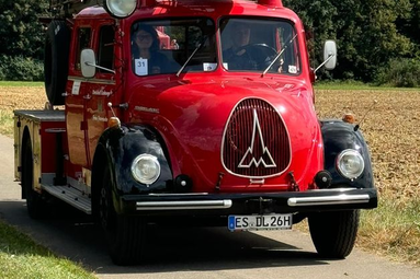 Historisches rotes Feuerwehrfahrzeug mit markanter Frontpartie und klassischem Magirus-Emblem, auf einer Landstraße entlang von Feldern unterwegs. | © Feuerwehr Oldtimerfreunde Esslingen 