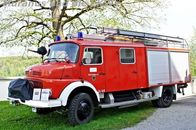 Historisches rotes Löschfahrzeug der Feuerwehr mit Blaulicht, Leiter und Dachausrüstung, auf einer Wiese unter Bäumen geparkt. | © Feuerwehr Oldtimerfreunde Esslingen 