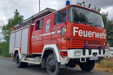 Rotes Feuerwehrfahrzeug der Freiwilligen Feuerwehr, geparkt am Straßenrand, mit Blaulicht, Leiter am Dach und markanter „Feuerwehr“-Beschriftung an der Front. | © Interessengemeinschaft "Emma" - LF 16 Nieder Wöllstadt e.V. 