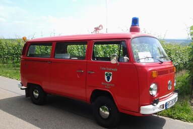 Historischer roter Feuerwehrmannschaftsbus der Freiwilligen Feuerwehr Weingarten, am Straßenrand in ländlicher Umgebung mit Blaulicht geparkt. | © Freiwillige Feuerwehr Weingarten/Baden 