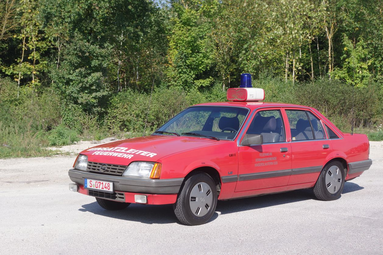 Rotes historisches Einsatzfahrzeug der Freiwilligen Feuerwehr, seitlich auf einem Parkplatz vor Bäumen stehend, mit Blaulicht am Dach. | © Förderverein Feuerwehr Wolpertshausen 