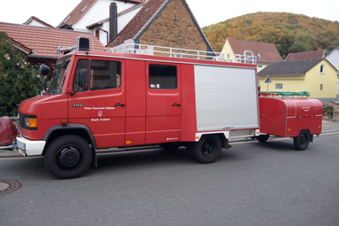 Historisches rotes Feuerwehrfahrzeug mit Anhänger, in einem Wohngebiet geparkt, mit seitlichen Geräteräumen und zusätzlicher Ausrüstung. | © Andreas Born 
