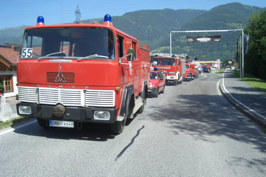 Kolonne historischer roter Feuerwehrfahrzeuge auf einer Straße in alpiner Umgebung, angeführt von einem klassischen Einsatzfahrzeug vor Bergkulisse. | © Förderverein Feuerwehr Wolpertshausen 