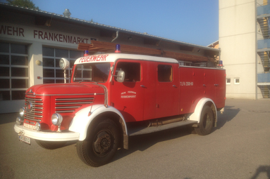 Historisches rotes Tanklöschfahrzeug der Feuerwehr Frankenmarkt, vor dem Feuerwehrhaus geparkt, mit klassischer Bauweise und Leiter auf dem Dach. | © Feuerwehr Oldtimer Verein Frankenmarkt 