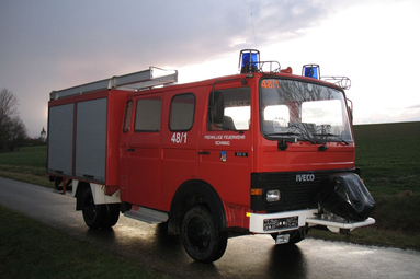 Historisches rotes Feuerwehrlöschfahrzeug der Freiwilligen Feuerwehr, auf einer Landstraße bei Abendstimmung mit Blaulicht und seitlichen Geräteräumen geparkt. | © Oldtimerfreunde Leuzendorf 