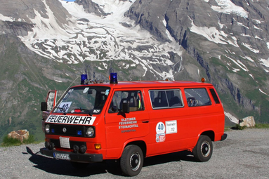 Historischer roter Feuerwehrmannschaftsbus vor hochalpiner Gebirgskulisse, auf einer Bergstraße geparkt, mit Blaulicht und markanter Feuerwehrbeschriftung. | © Andreas Kossak 